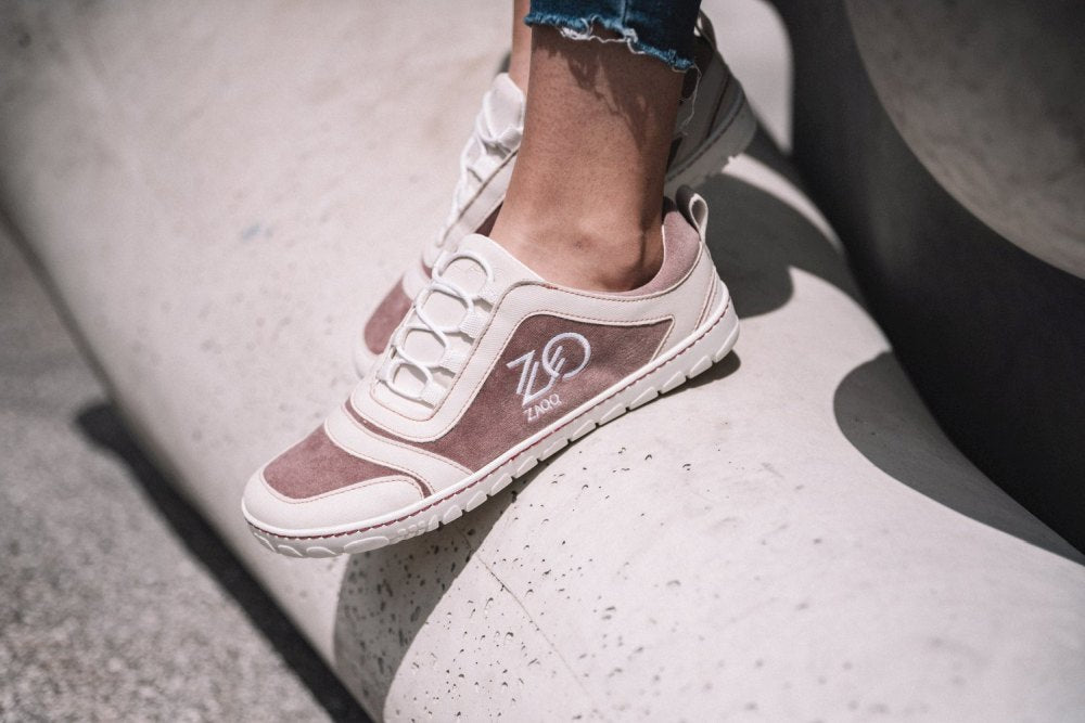 Eine Person, die modische beige-weiße SQILL Rose-Sneaker von ZAQQ trägt, mit Z&D-Stickerei an der Seite und aus veganer Mikrofaser gefertigt, steht auf einer glatten, geschwungenen Betonoberfläche. Die Sneaker haben ein lässiges Design, das perfekt zu Jeans mit ausgefranstem Saum passt.