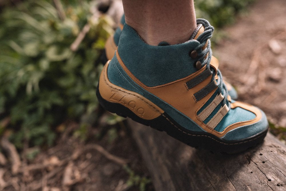 Nahaufnahme einer Person, die die wasserdichten Wanderstiefel SQOUT Blue Orange von ZAQQ trägt. Sie sind in den Farben Blaugrün und Hellbraun gehalten, haben schwarze Sohlen und ein Logo an der Seite. Diese Stiefel sind sowohl atmungsaktiv als auch wasserdicht. Die Person steht selbstbewusst auf einem Baumstamm, im Hintergrund ist verschwommenes grünes Laub zu sehen.