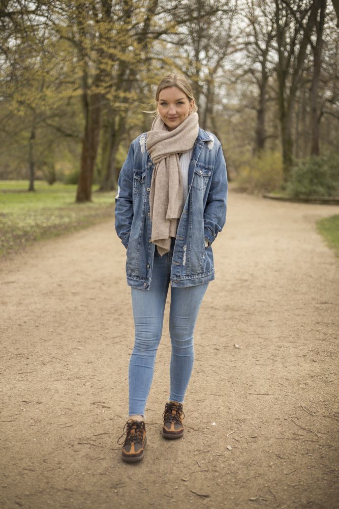 Jemand läuft auf einem Feldweg im Park, umgeben von Bäumen. Er trägt eine Jeansjacke, hellblaue Jeans, einen beigen Schal und robuste, braune wasserdichte SQOUT-Schuhe von ZAQQ mit TERRA-Sohlen. Er läuft selbstbewusst unter einem bedeckten Himmel, der einen kühlen Tag ankündigt.