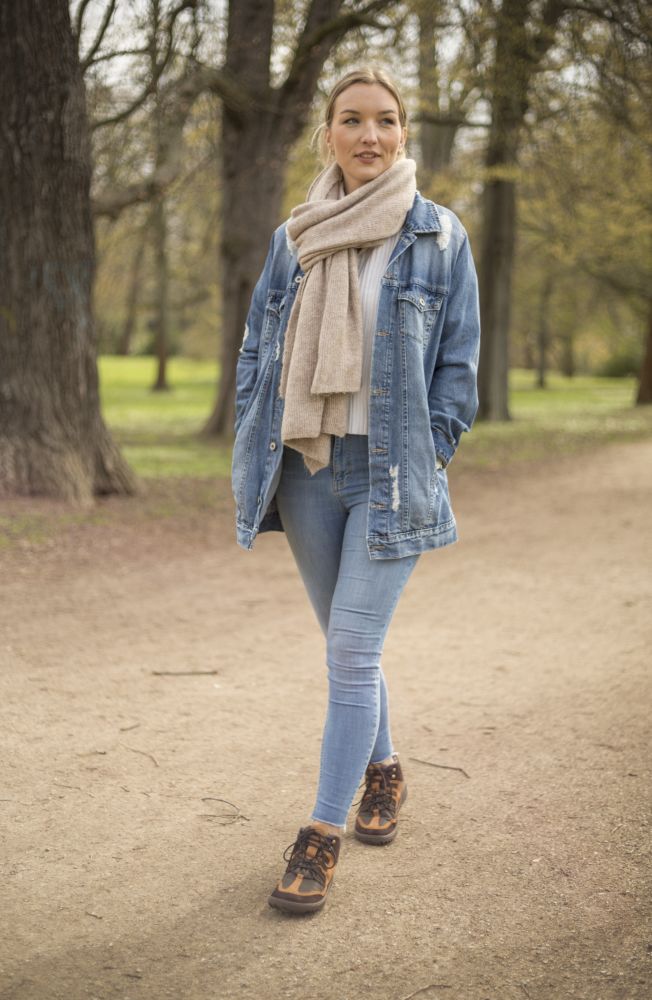 Eine Person schlendert auf einem Feldweg durch einen von Bäumen umgebenen Park. Sie trägt eine Jeansjacke, einen beigen Schal, blaue Jeans und die zuverlässigen braunen wasserdichten ZAQQ SQOUT-Stiefel für jedes Gelände. Ihre Haare sind zu einem Pferdeschwanz zurückgebunden. Die Kulisse ist üppig grün.