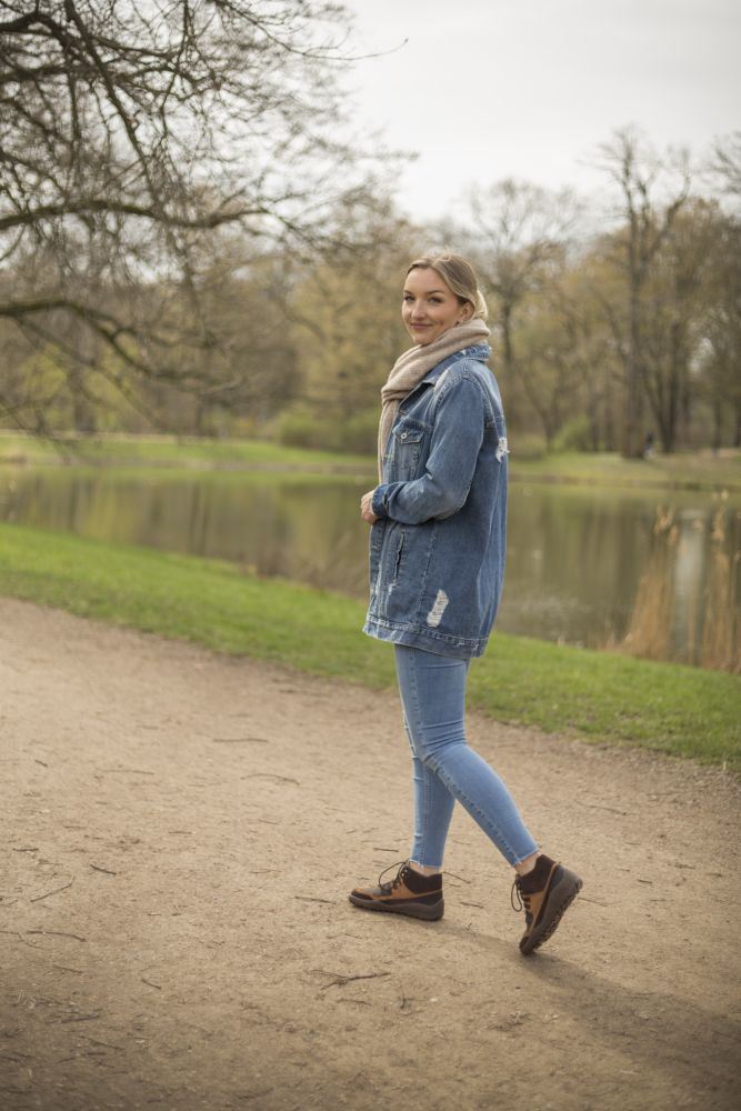 Eine Person in Jeansjacke und Jeans schlendert einen Feldweg neben einem Teich entlang. Sie trägt die SQOUT Brown Waterproof-Schuhe von ZAQQ, die mit ihrer TERRA-Sohle Halt auf dem Boden bieten. Die kahlen Bäume und das grüne Gras lassen auf den frühen Frühling schließen, und der bedeckte Himmel trägt zur ruhigen, natürlichen Atmosphäre bei.