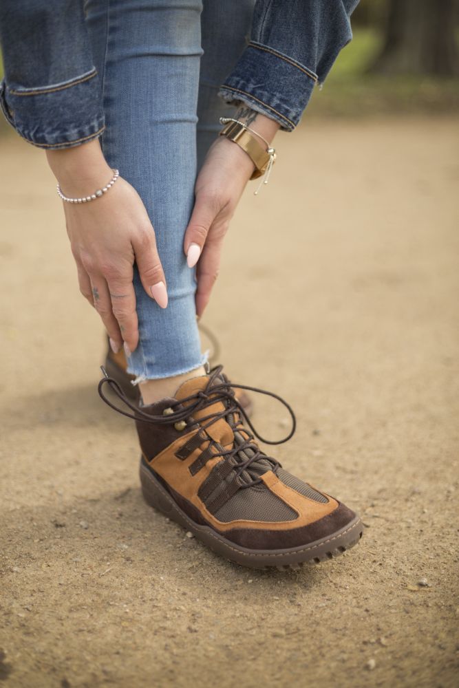 In Bluejeans und Jeansjacke über einem braunen Oberteil zieht eine Person auf dem Feldweg ihren braunen wasserdichten SQOUT-Stiefel mit schwarzen Schnürsenkeln zurecht. Sie präsentiert Perlen- und Goldarmbänder sowie manikürte Nägel, während die ZAQQ TERRA-Sohle durch ihre Velourslederverarbeitung für einen stabilen Halt sorgt.