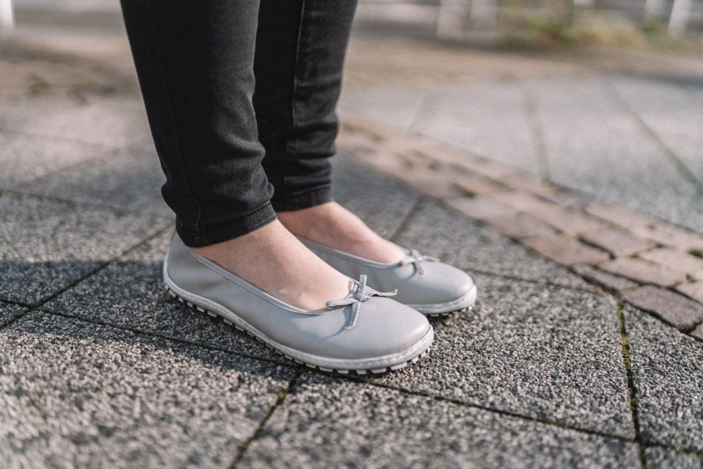 Eine Person in schwarzen Hosen und hellgrauen SUQAR-Ballerinas mit kleinen Schleifen steht anmutig auf einem strukturierten Steinpflaster. Das Design ihrer ZAQQ-Barfußschuhe sorgt für Komfort und zugleich für Eleganz.