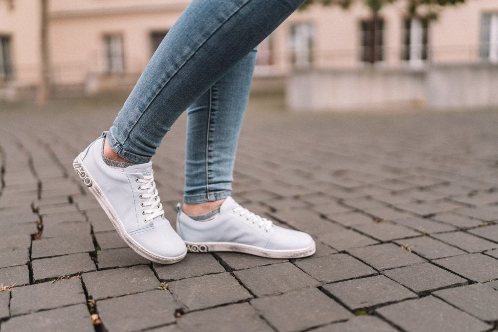 Eine Person trägt die TAQQ Nappa Lightblue Sneaker von ZAQQ mit flexibler Sohle und betritt in Jeans einen Kopfsteinpflasterweg. Der Hintergrund ist leicht verschwommen und zeigt ein Gebäude und einige Bäume. Die Schuhe haben weiße Schnürsenkel und eine Gummisohle, was subtil auf den Komfort des Barfußschuh-Designs hinweist.