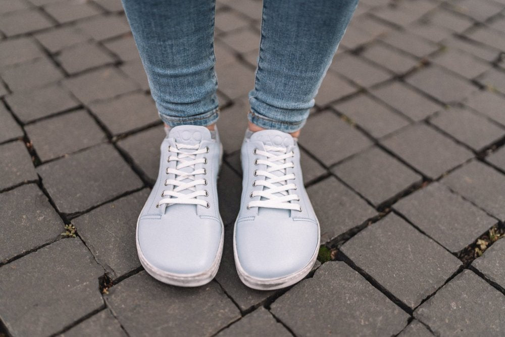 Nahaufnahme einer Person, die die TAQQ Nappa Lightblue Sneaker von ZAQQ aus hochwertigem Nappaleder trägt. Diese hellblauen Barfußschuhe sind stilvoll geschnürt und mit Blue Jeans kombiniert. Sie stehen auf einer Pflastersteinoberfläche und zeigen ihre leicht abgenutzten, aber flexiblen Sohlen.