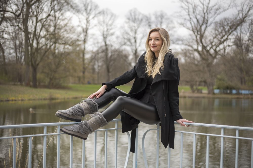 Eine Frau in schwarzem Mantel und Leggings sitzt an einem bewölkten Tag auf einem Metallgeländer an einem See, umgeben von kahlen Bäumen. Sie trägt graue TORQ Winterstiefel von ZAQQ, die ein Barfußgefühl vermitteln, und lächelt, während sie in die Kamera blickt.