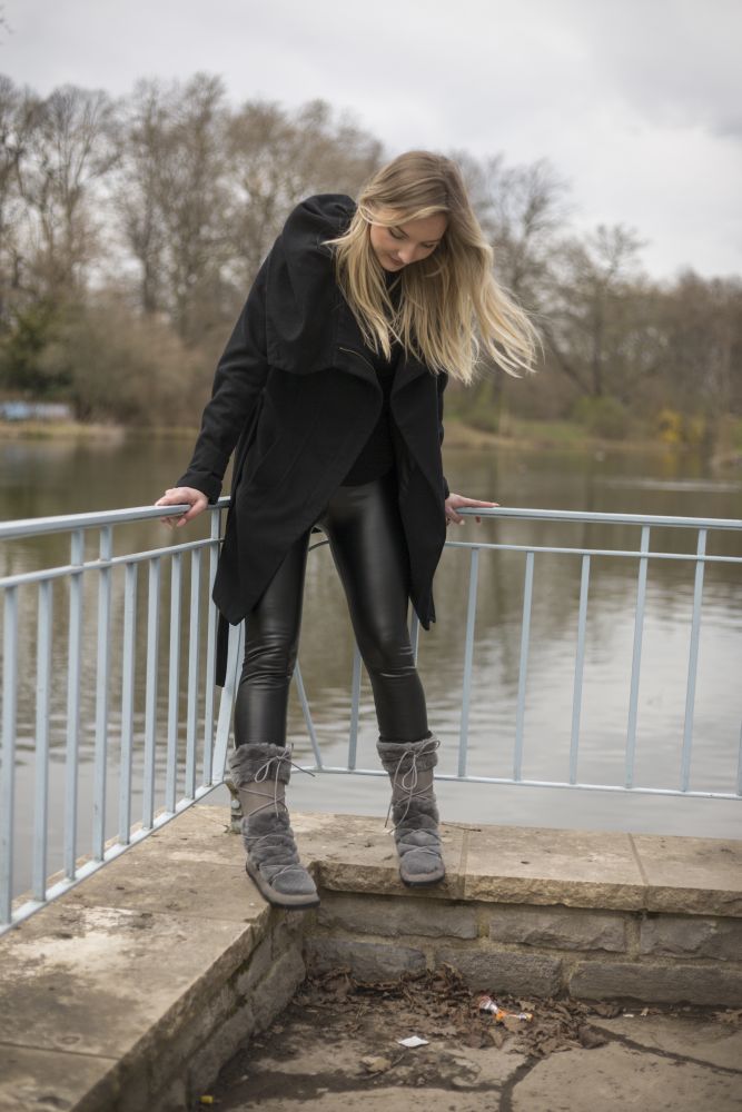 Eine Person steht auf einer Steinplattform an einem See und hält sich an einem Metallgeländer fest. Sie trägt einen schwarzen Mantel, schwarze Lederhosen und graue ZAQQ TORQ Winterstiefel, die ein Barfußgefühl vermitteln. Im Hintergrund sind kahle Bäume und ein bewölkter Himmel zu sehen.