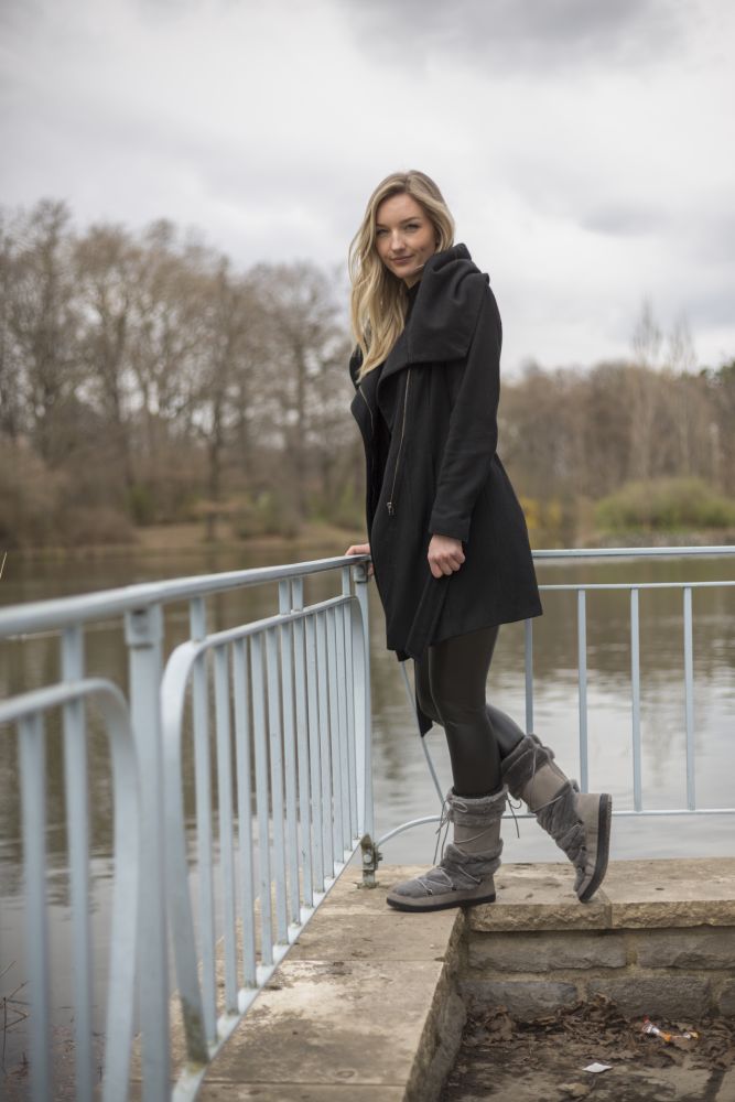 Eine Person in einem schwarzen Mantel und grauen TORQ Winterstiefeln von ZAQQ steht auf einem kleinen Pier am Wasser. Im Hintergrund sind kahle Bäume unter einem bewölkten Himmel zu sehen.