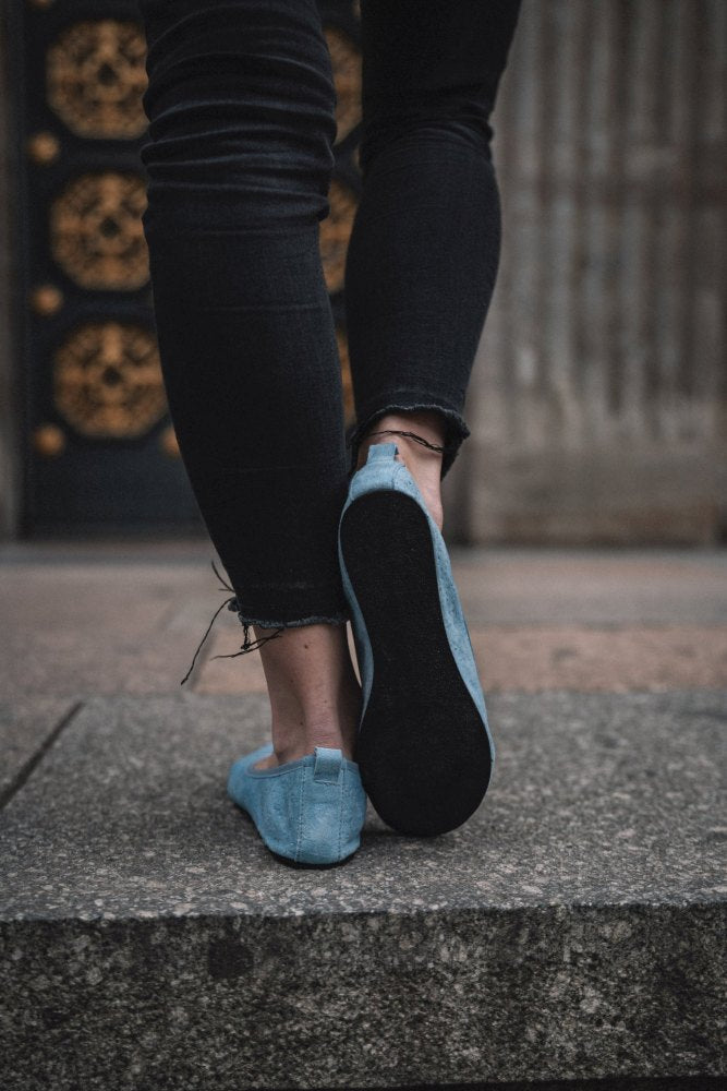 Eine Person trägt die Slipper TWIST Light Blue von ZAQQ und zerrissene schwarze Jeans und betritt eine Steinfläche. Im Hintergrund ist eine dekorative, dunkel gefärbte Tür mit goldenen Details zu sehen, die schicken Sommerlooks einen Hauch von Eleganz verleiht.