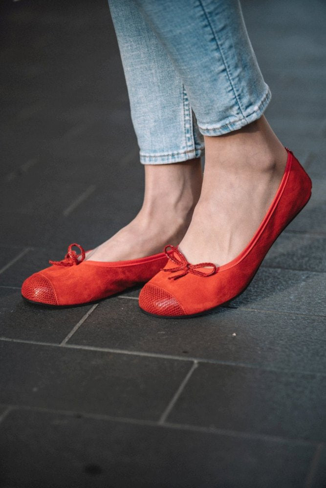 Eine Nahaufnahme zeigt jemanden, der die TWIST Velours Red Ballerinas von ZAQQ trägt. Diese stylischen roten Schuhe haben vorne eine kleine Schleife und glitzernde Zehenpartien. Die Person steht auf einem dunklen Fliesenboden und trägt dazu hellblaue Jeans.