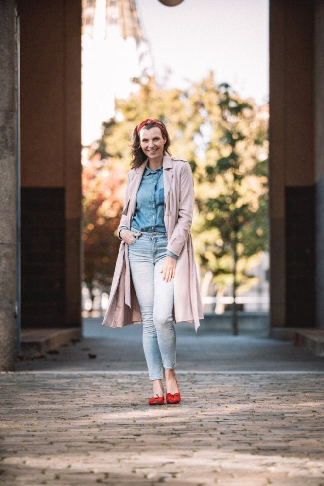 Eine Frau steht auf einem Kopfsteinpflasterweg und trägt einen langen rosa Mantel, ein Jeanshemd und Jeans. Sie lächelt mit einem roten Stirnband und passenden TWIST Velours-Schuhen von ZAQQ. Sonnenlicht fällt durch die Bäume im Hintergrund und schafft eine warme und einladende Atmosphäre.