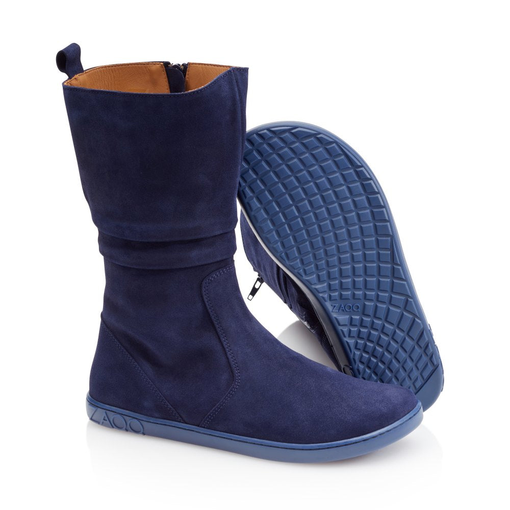 Ein Paar QOM Blue Wildlederstiefel von ZAQQ in einem dunklen Blauton hat eine flache Sohle und ein halbhohes Design. Ein Stiefel steht aufrecht und zeigt einen sichtbaren Reißverschluss, und der andere liegt auf der Seite, sodass die strukturierte ZAQQ-Barfußsohle sichtbar ist. Das Innenfutter ist hellbraun und das ZAQQ-Branding ist in die Sohle eingeprägt.