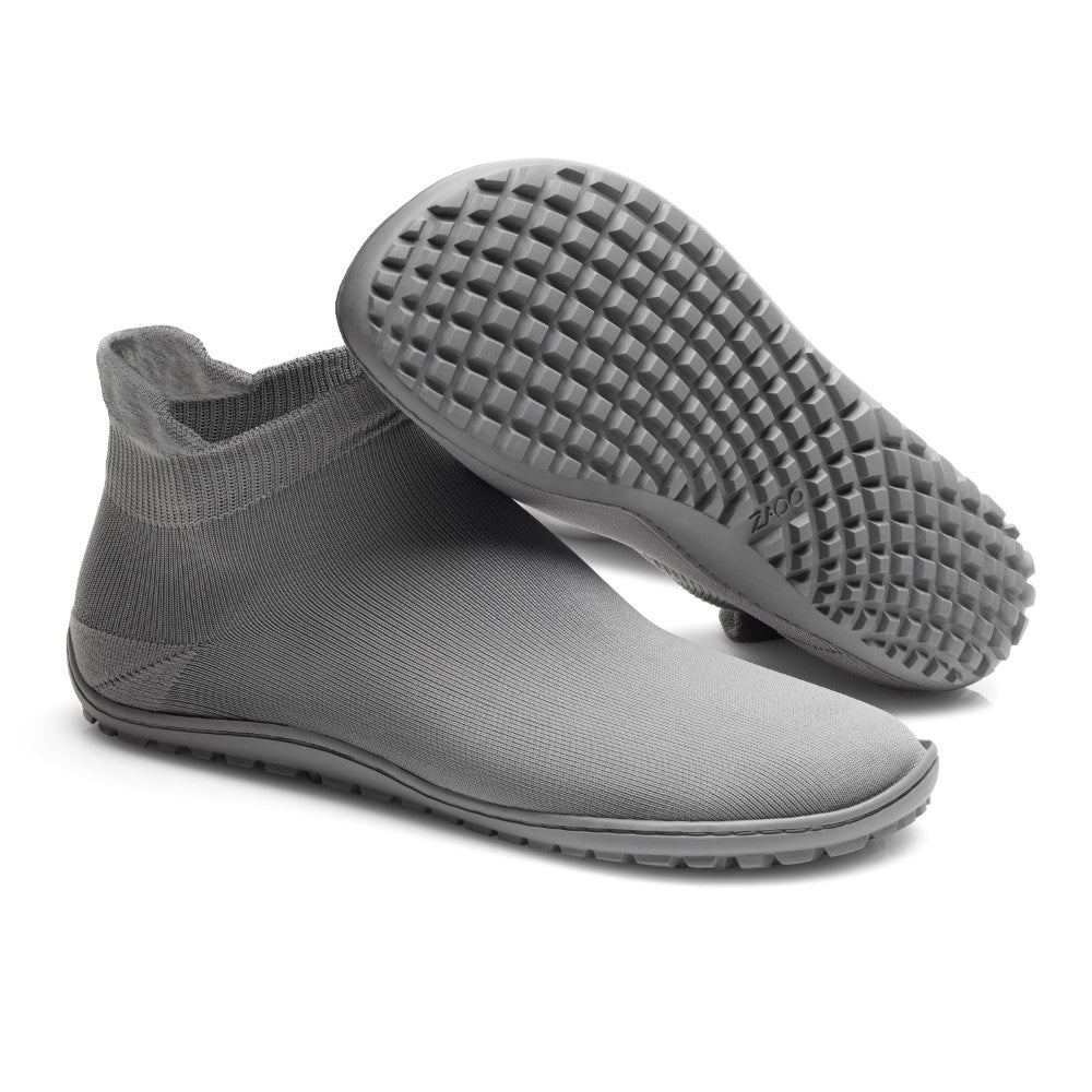 Wir präsentieren den SOQQ Grey von ZAQQ: Dieses Paar stylischer knöchelhoher Schuhe verfügt über strukturierte Gummisohlen mit einem markanten Rutschmuster. Ein Schuh steht aufrecht, während der andere auf der Seite liegt, wodurch das strapazierfähige und flexible Strickgewebe in einem eleganten Grauton hervorgehoben wird.